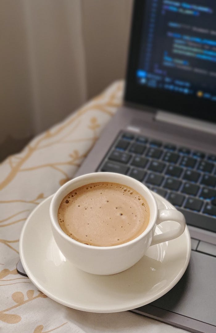 Cup of frothy coffee on a laptop keyboard with coding screen, cozy indoor setting.